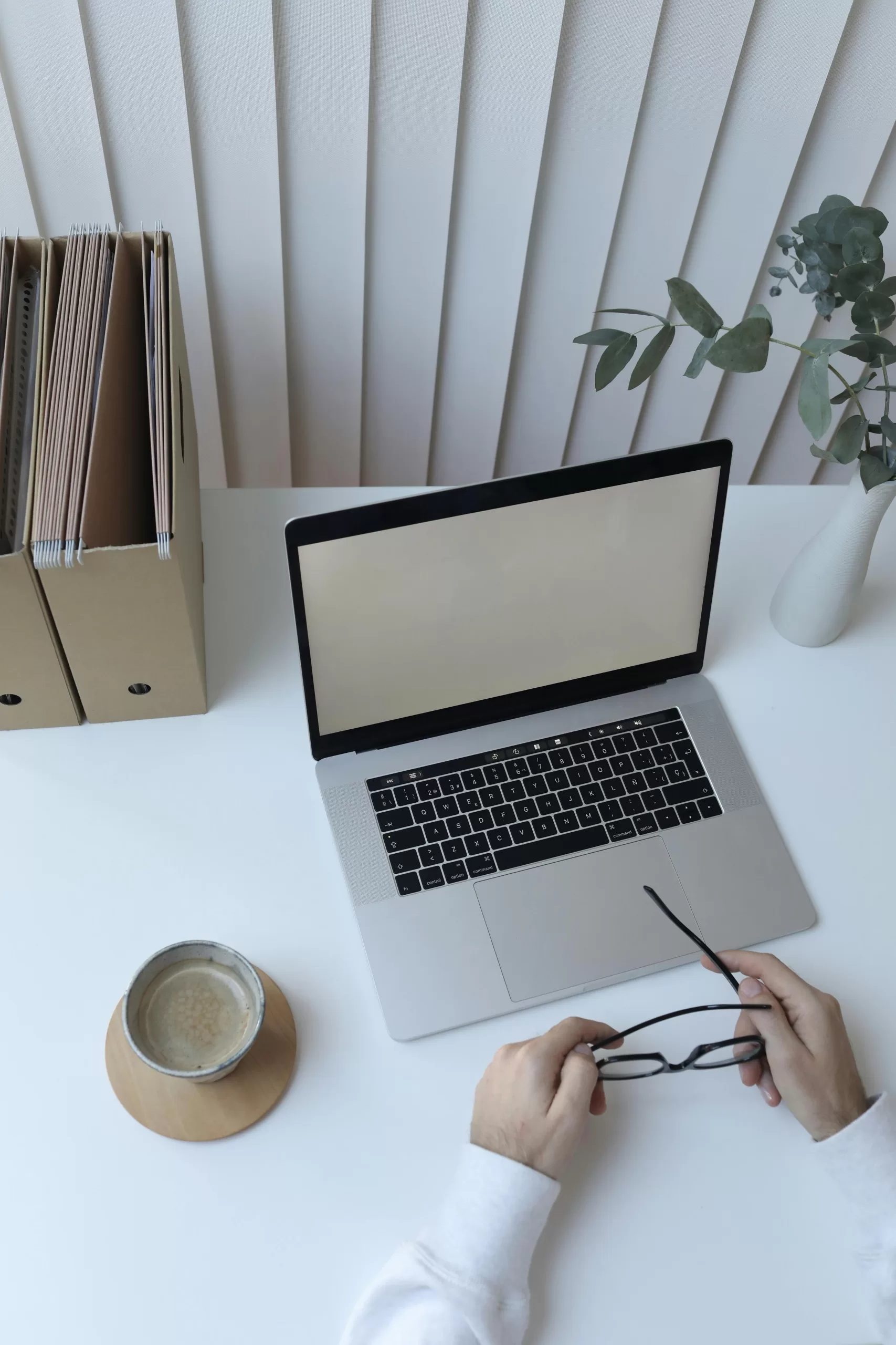 Workspace setup with a laptop, a pair of hands holding glasses, a cup of coffee, file organizers, and a vase with green leaves.