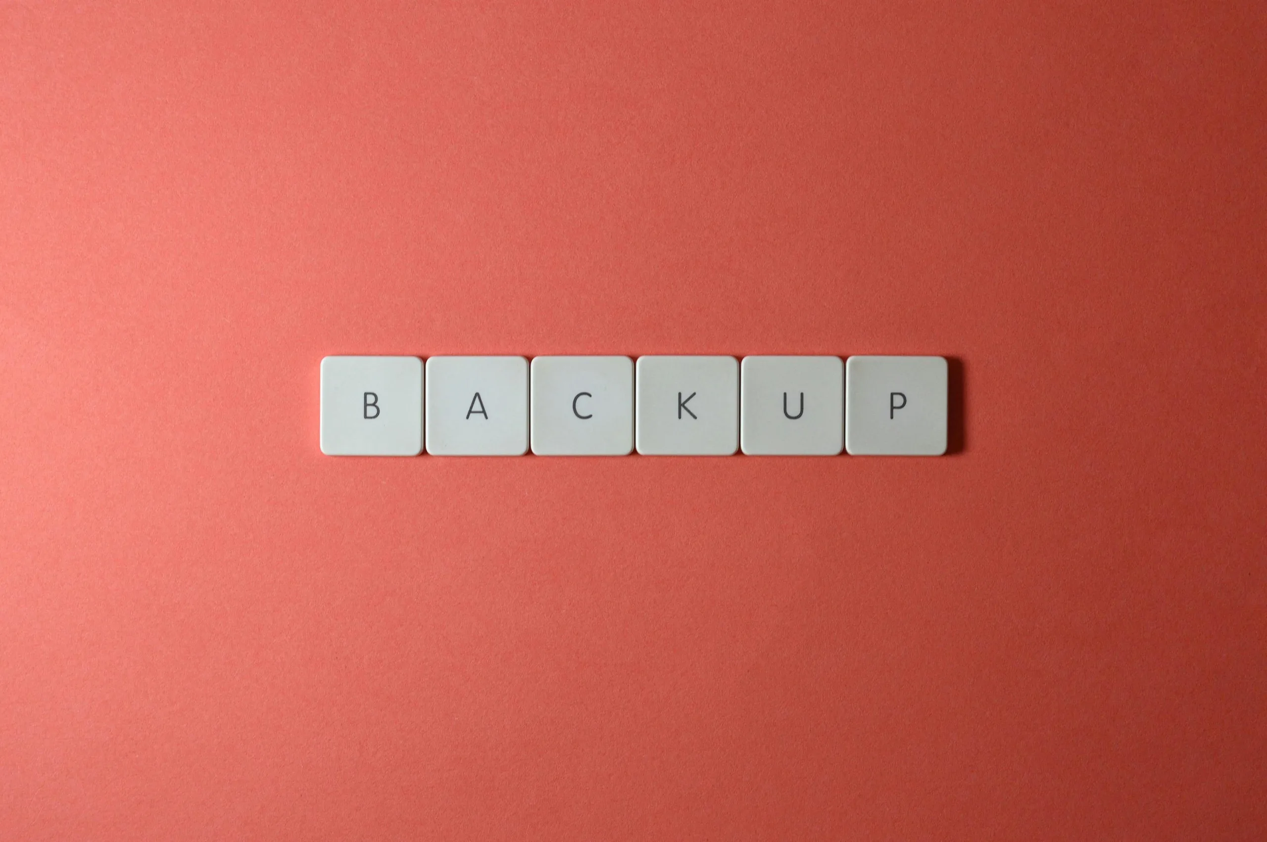 White keyboard tiles spelling “BACKUP” on a flat coral-coloured background.