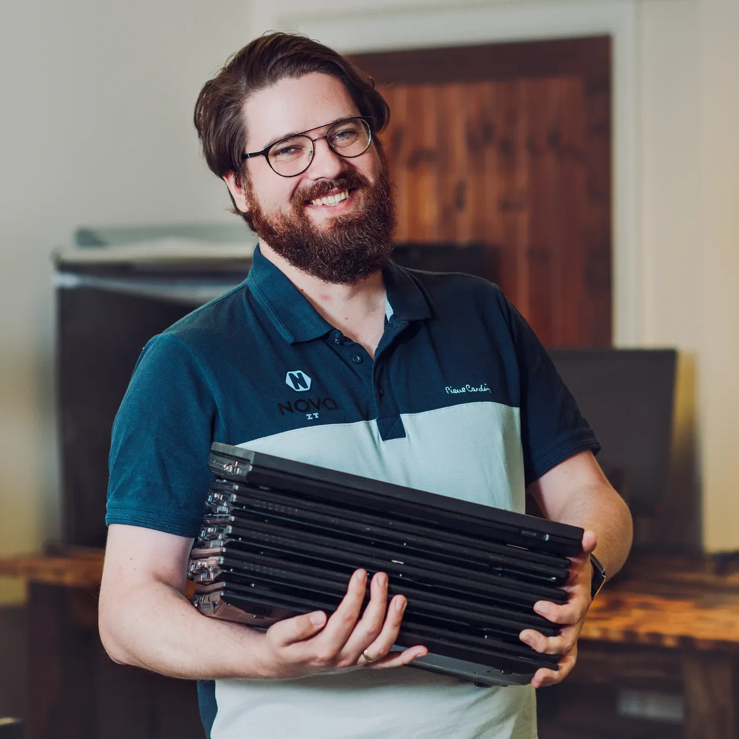 Szymon Dymek smiling and holding a stack of laptops while wearing a Novo IT branded polo shirt.