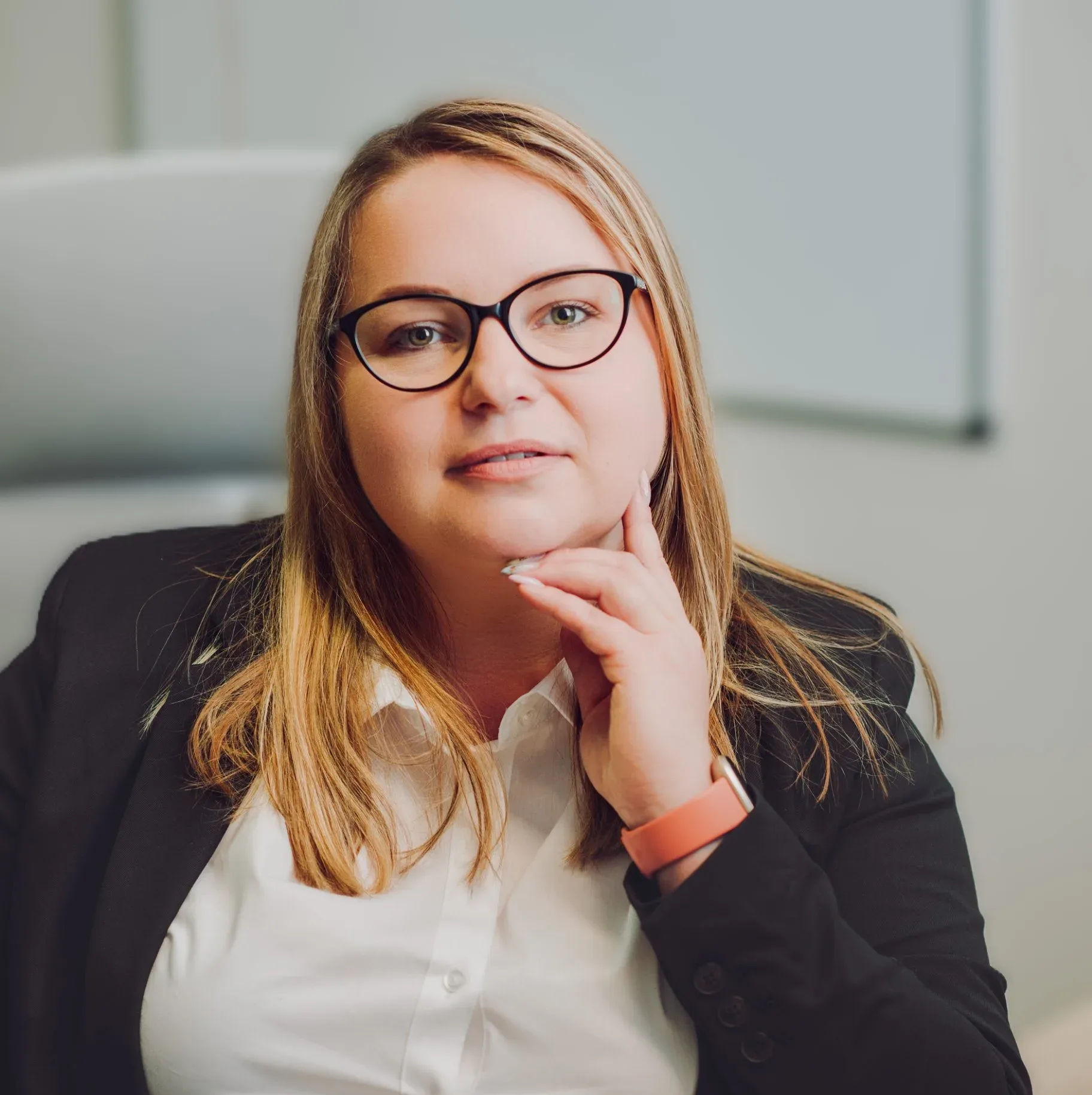 Anita Dymek sitting at a desk, wearing glasses and a black blazer, looking confidently into the camera with hand resting on chin.