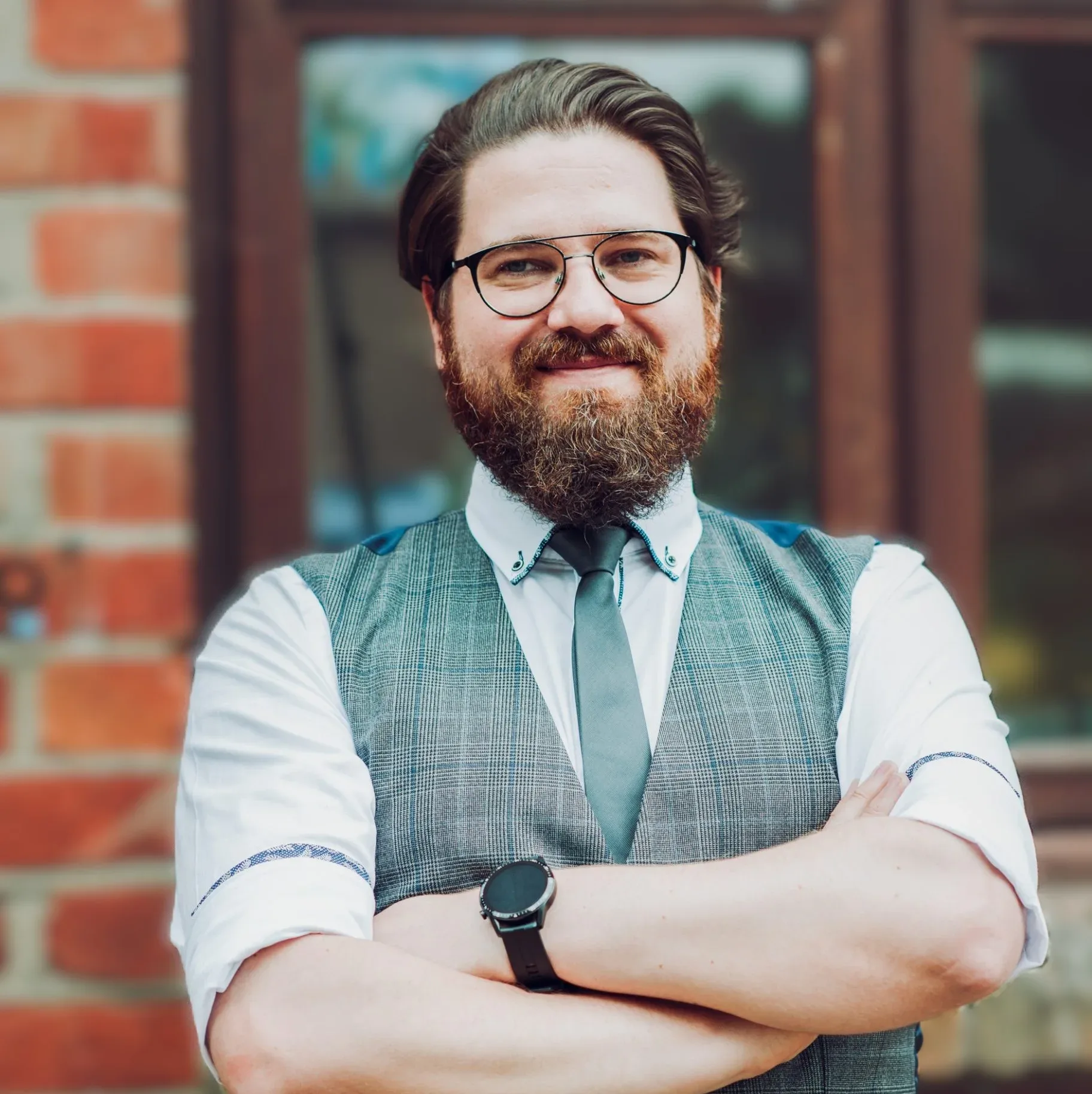 Szymon Dymek standing outside against a brick wall, smiling with arms crossed, wearing glasses, a grey waistcoat, and tie.