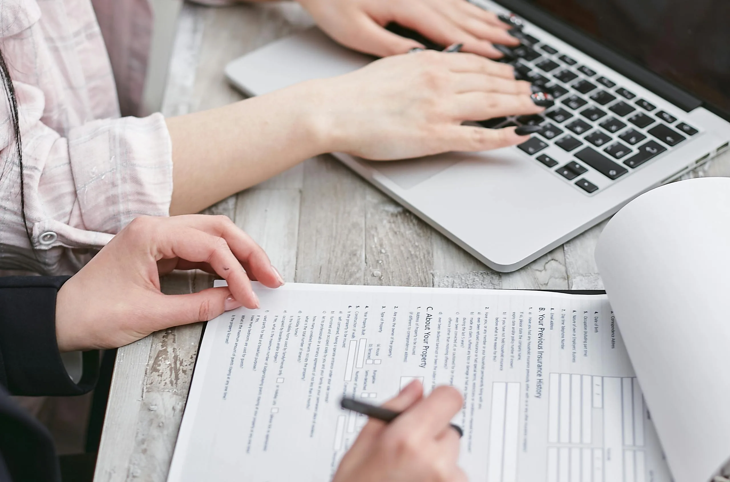 Hands filling out cyber insurance policies beside a laptop user—representing digital and paper-based workflows.