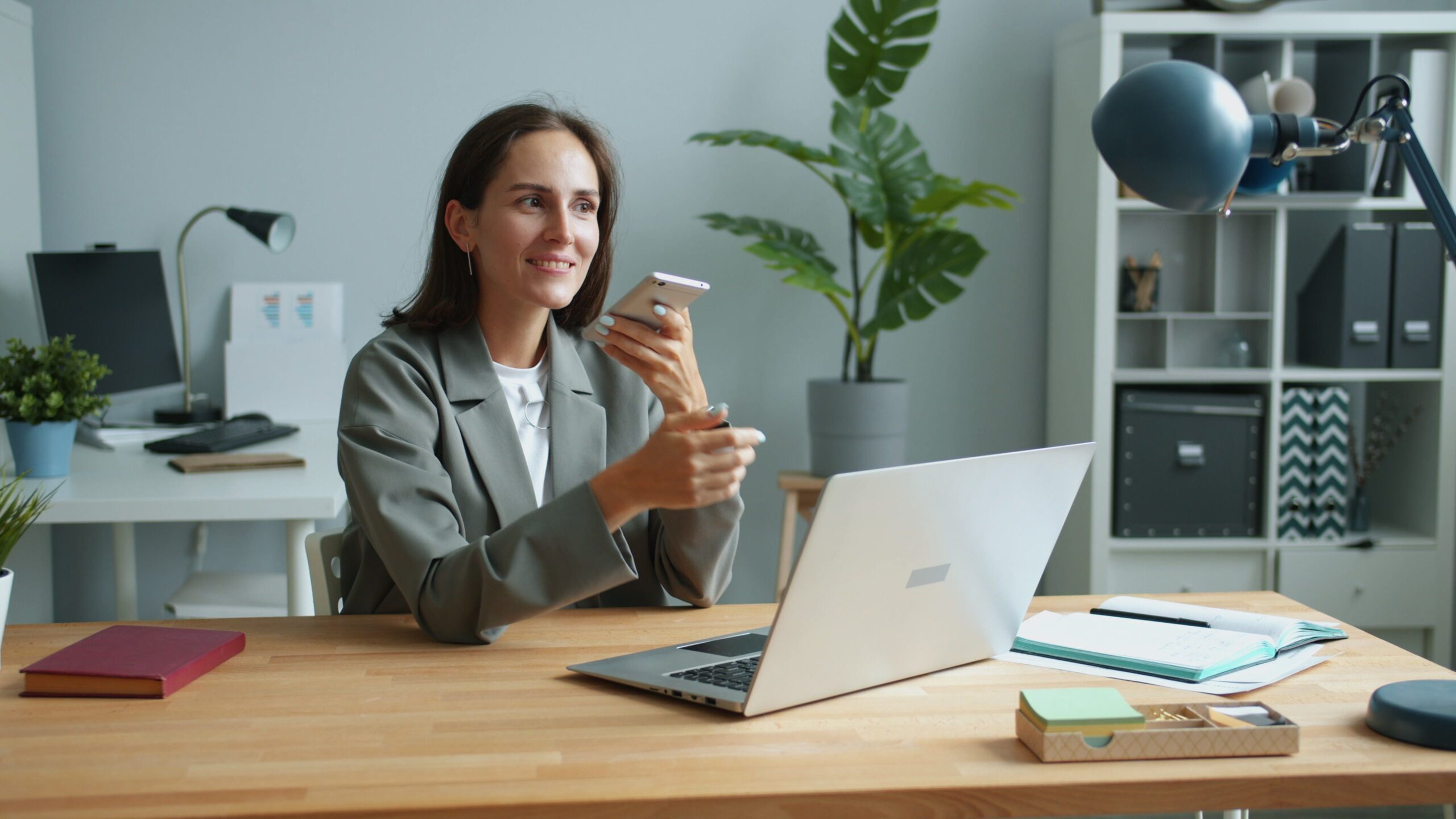 A smiling woman sitting at a desk with an open laptop, holding a smartphone near her face to speak or listen.