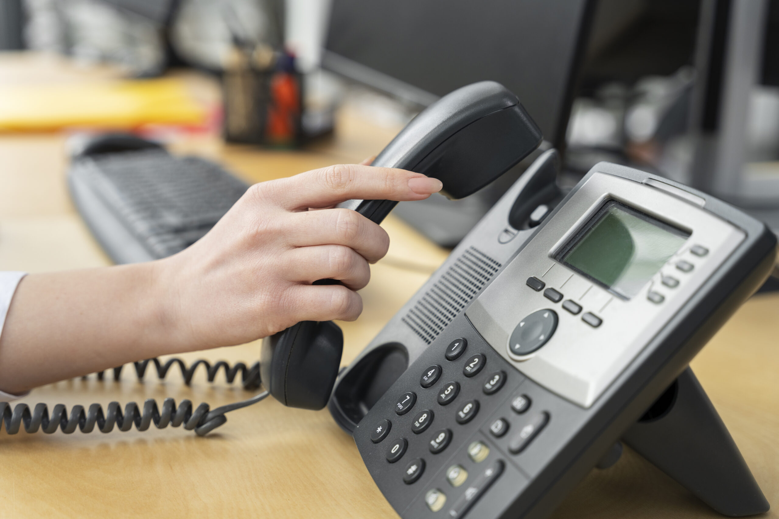 Close-up of a person's hand lifting the handset of a grey office desk telephone on a wooden desk.